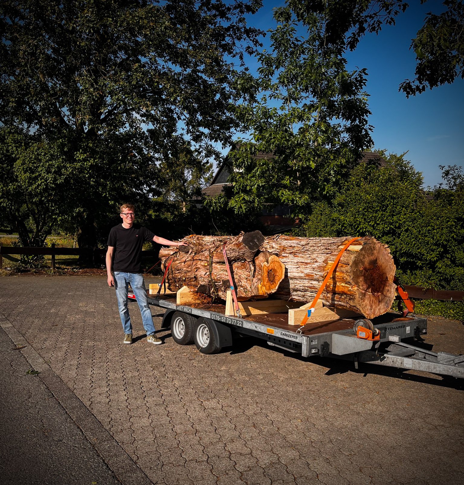 Leo_Weyherter_steht_neben_einem_grossen_Holzstamm__den_er_fuer_Schorn___Groh_erworben_hat